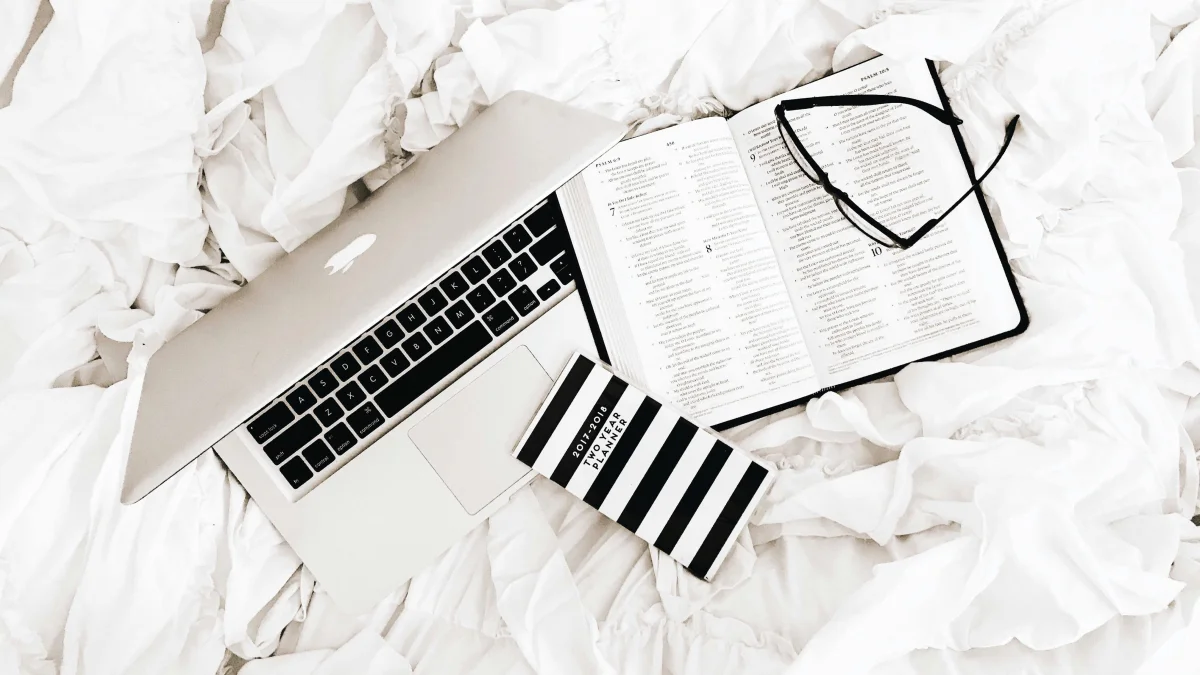 A book, notebook and glasses laying on a white bed
